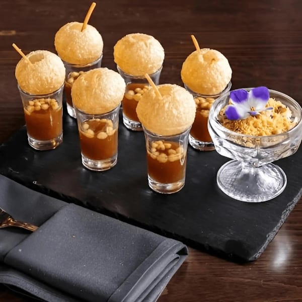 Artha's pani puri shots, with crispy puris atop glasses of spiced water and chickpeas, on a black tray.