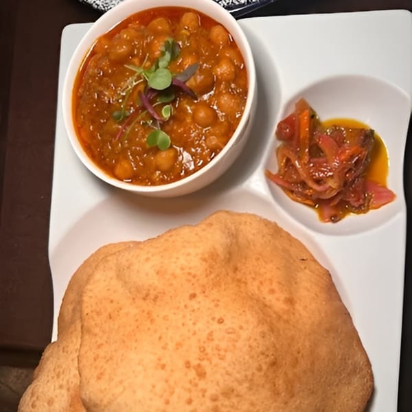 Chana masala with microgreens, pickled onions, and a large puri bread on a white plate.