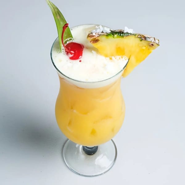 A vibrant yellow cocktail with a frothy top, garnished with a cherry, pineapple slice, and green leaf.