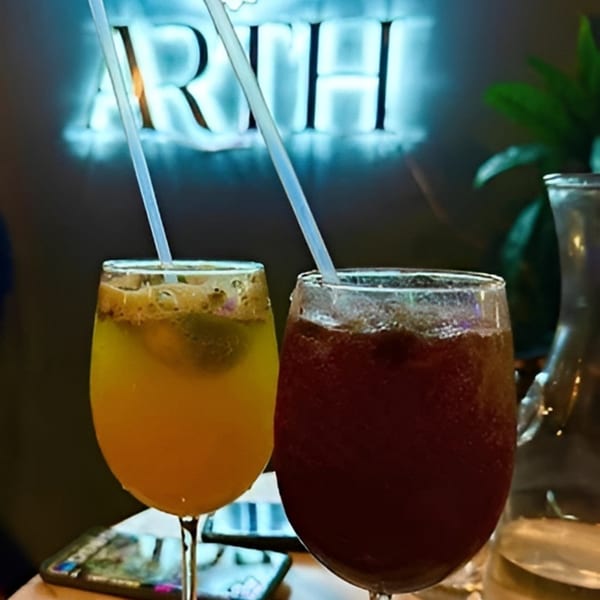 Two cocktails, one yellow and one red, with straws, in front of a neon "ARTHA" sign.