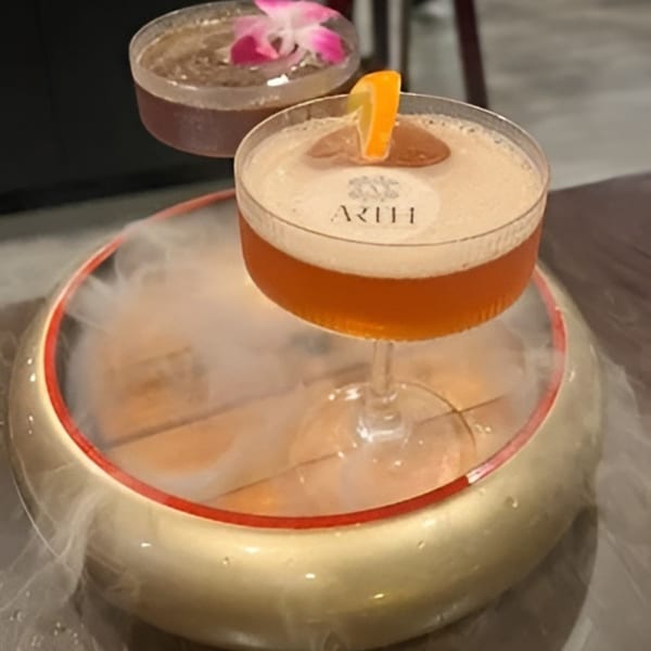 Two cocktails, one with an orange slice and the other with a pink flower, on a smoky golden bowl.